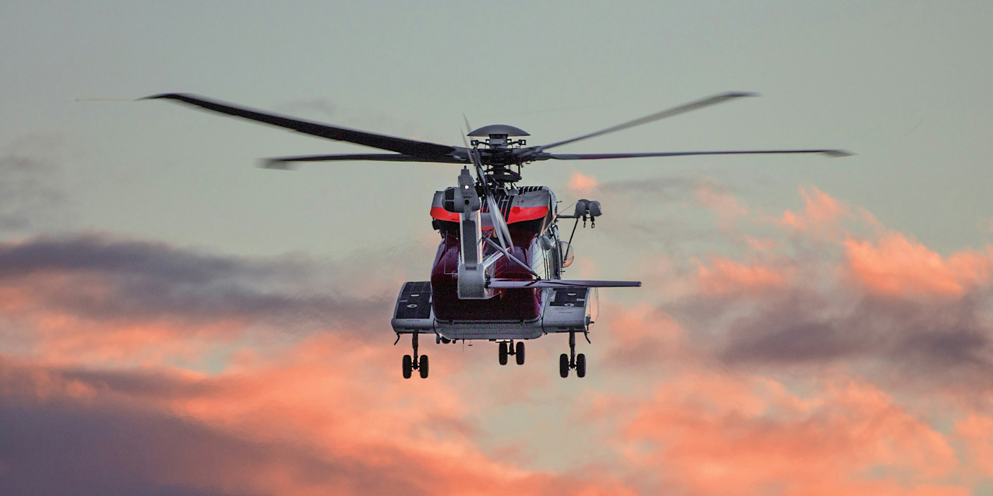 Et rødt og hvitt redningshelikopter sett bakfra i lufta med landingshjulene ute. Hovedrotoren er i bevegelse og fremstår uskarp. Helikopteret flyr mot en himmel med dramatiske skyer i varme rosa og oransje toner ved soloppgang eller solnedgang.