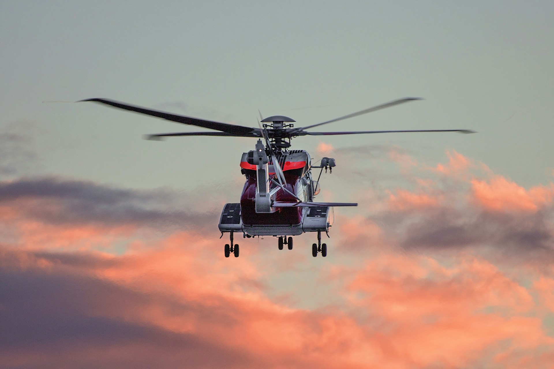 Et rødt og hvitt redningshelikopter sett bakfra i lufta med landingshjulene ute. Hovedrotoren er i bevegelse og fremstår uskarp. Helikopteret flyr mot en himmel med dramatiske skyer i varme rosa og oransje toner ved soloppgang eller solnedgang.